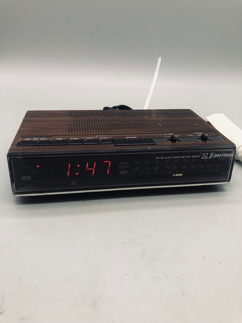 以下が含まれることがあります： 1:47 を表示するデジタルディスプレイを搭載した、茶色と黒の Emerson クロックラジオ。ラジオには内蔵アラームクロックとバッテリーバックアップ機能があります。ラジオは黒と銀のフェイスで、白いアンテナが付いています。