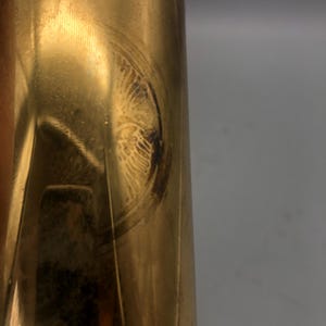 May include: Close-up of a brass-coloured, cylindrical object with a circular, etched design. The design appears to be an emblem or decorative element, possibly vintage. The surface shows signs of wear and age, with some discolouration.