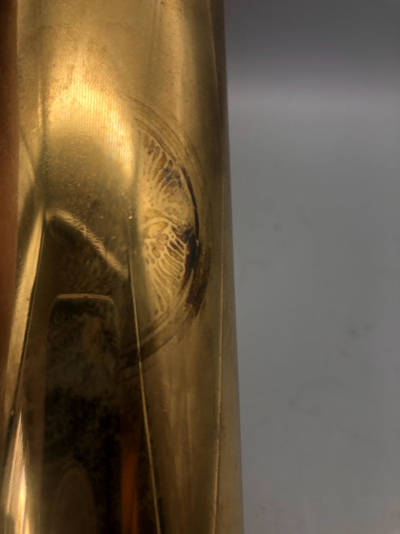 May include: Close-up of a brass-coloured, cylindrical object with a circular, etched design. The design appears to be an emblem or decorative element, possibly vintage. The surface shows signs of wear and age, with some discolouration.