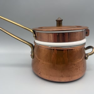 May include: Copper double boiler with a lid and brass handles. The pot has a white band around the middle and a small handle on the side. The lid has a small brass knob.