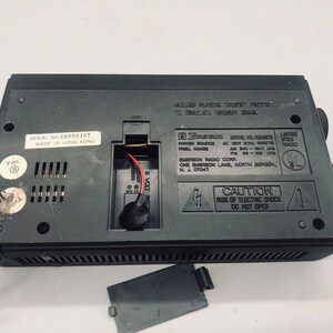 以下が含まれることがあります： 黒いエマーソンラジオの背面。シリアル番号、モデル番号、電源に関する情報が記載されています。ラジオにはバッテリーコンパートメントと、「感電の恐れあり。開けないでください。」という注意書きのラベルが付いています。