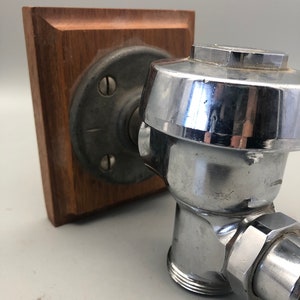 May include: A chrome-plated toilet tank valve with a wooden mounting plate. The valve has a round, chrome-plated cap and a threaded connection.