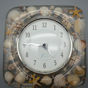 May include: Square clock with a white face, silver trim, and black numbers. The clock is encased in clear resin, embedded with seashells and starfish. The clock face reads "Shore Shells".