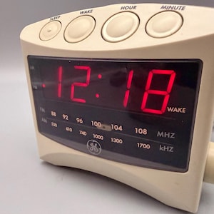 May include: A beige GE digital alarm clock radio with a black display panel showing the red LED time 12:18. The top features round buttons labeled SLEEP, WAKE, HOUR, and MINUTE. The front panel displays AM/FM radio frequencies and a WAKE indicator.