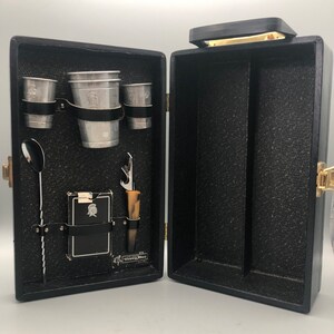 May include: Black leather bar set with a silver cocktail shaker, two silver cups, a silver jigger, a silver spoon, a bottle opener, and a card with a logo. The set is open and the interior is lined with black felt.