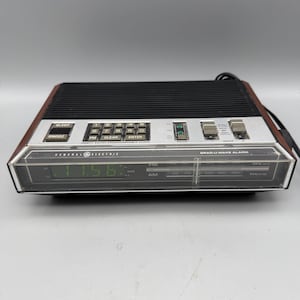 May include: A General Electric Grad-U-Wake alarm clock with a black top speaker grille, a silver control panel featuring a numeric keypad, and a digital display showing 11:56. The unit has a wood-grain side finish and an analog radio tuner.