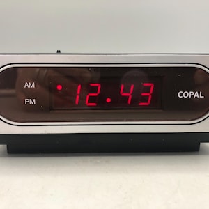 May include: A vintage digital alarm clock with a silver frame and a black base. The time, 12:43, is displayed in bright red numerals. The brand name "COPAL" is visible on the right side of the display.