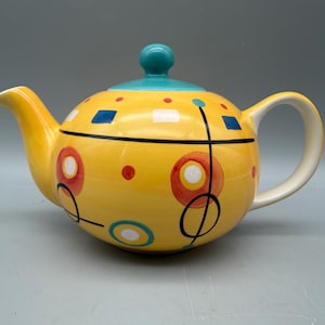 May include: A yellow ceramic teapot featuring a teal lid and a colorful pattern of red circles, blue squares, and thin black lines. The teapot has a curved handle and a spout, with a smooth, glossy finish and a rounded body shape.