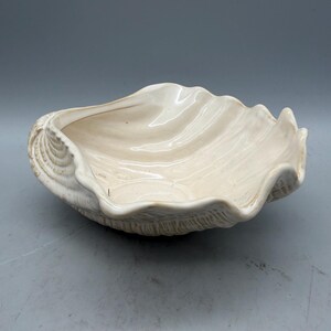 May include: A cream-colored, ceramic seashell-shaped bowl. The bowl has a wavy, textured rim and a smooth interior. The exterior has detailed shell patterns. This decorative bowl is suitable for coastal or nautical-themed decor.