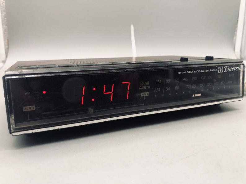 以下が含まれることがあります： 赤いデジタルディスプレイに1:47が表示された、黒いエマーソンFM-AMクロックラジオ。ラジオにはバッテリーバックアップ機能とデュアルアラーム設定があります。