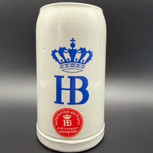 May include: A cylindrical light gray stoneware beer mug featuring a blue crown logo above the letters HB. Below the logo is a circular red sticker with white text reading Hofbräuhaus am Platzl, HB, and H.Glanegger Hofbräuhauswirt.