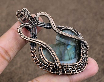 Puede incluir: Un colgante de labradorita en forma de corazón, engastado en un intrincado entrelazado de alambre de cobre oxidado. El trabajo de alambre presenta un patrón complejo de bucles y espirales que rodean la piedra, la cual muestra destellos iridiscentes azules y verdes.