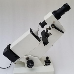 Puede incluir: Un lensómetro blanco y negro con una lente negra y una perilla negra en el lateral. El lensómetro está sobre una superficie blanca.