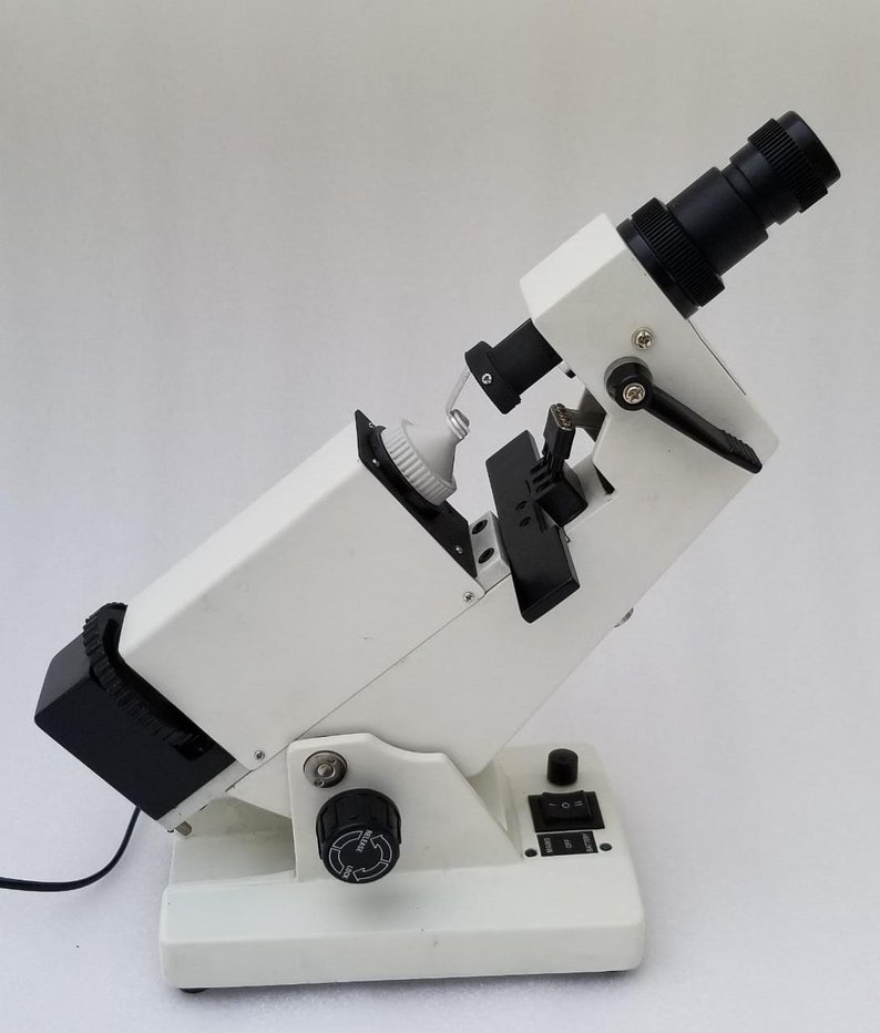 May include: A white and black lensometer with a black lens and a black knob on the side. The lensometer is on a white surface.