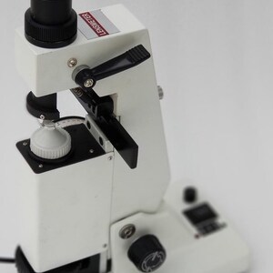 May include: A white lensmeter with a black lens and a black knob. The lensmeter has a black lever and a white dial with black numbers. The lensmeter is on a white surface.
