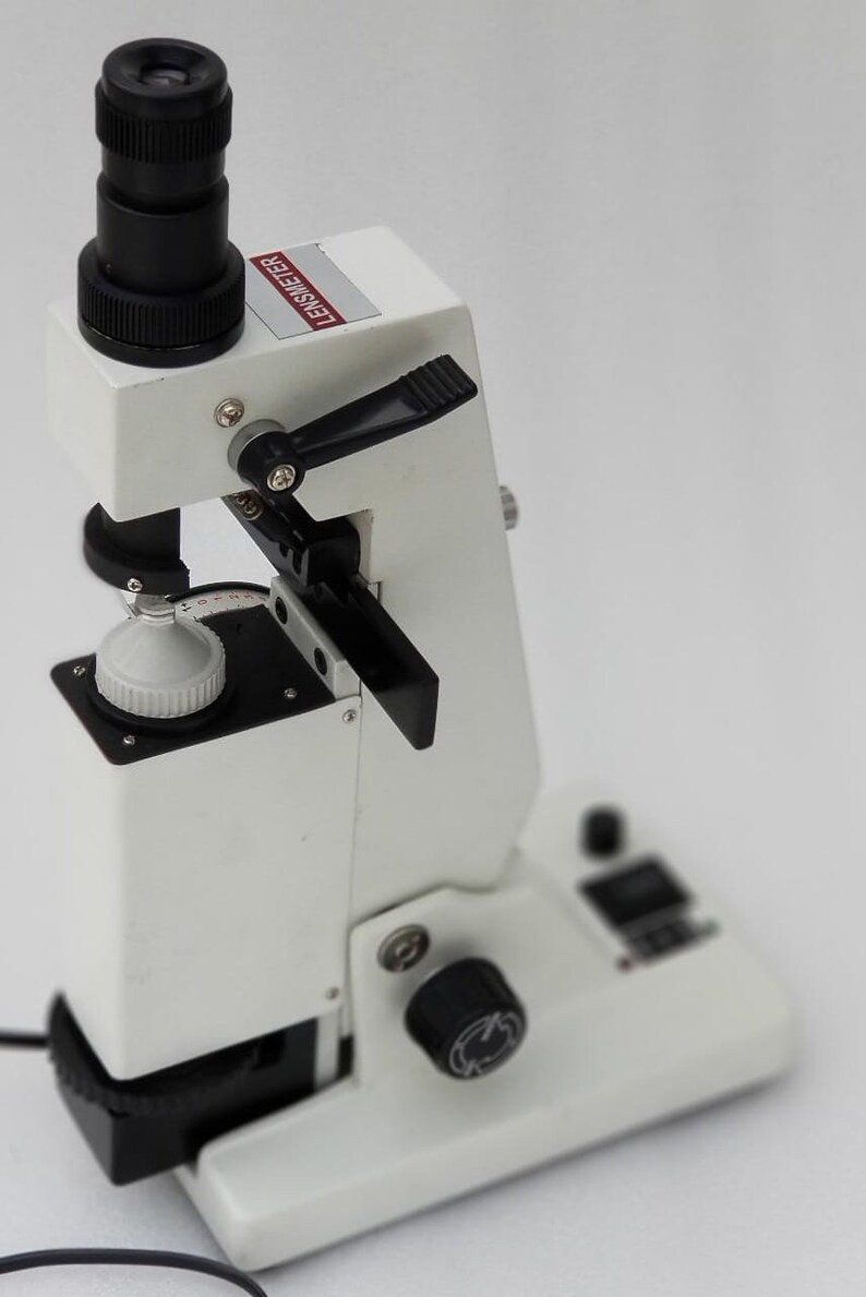 May include: A white lensmeter with a black lens and a black knob. The lensmeter has a black lever and a white dial with black numbers. The lensmeter is on a white surface.