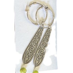 May include: Silver-toned hoop earrings with long, ornate pendants featuring a Celtic knot design. Each earring has a small, round, light green bead at the bottom. The earrings are displayed on a white surface.