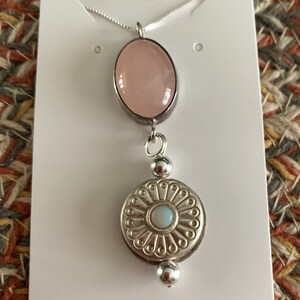 May include: A silver necklace with a pendant featuring a large, oval, pink gemstone above a detailed, circular, silver pendant with a smaller, round, light blue stone in the center. The necklace is displayed on a white card.