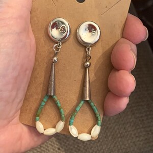 May include: Silver-toned dangle earrings with turquoise and white bead accents. The earrings feature a silver stud, a cone-shaped element, and a teardrop design made of turquoise and white beads. The earrings are displayed on a brown card.