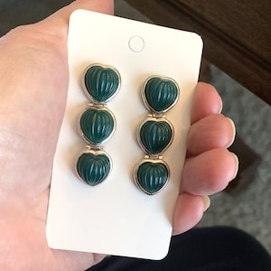 May include: A pair of dangling earrings featuring three heart-shaped, dark green pendants with a ribbed texture. The pendants are connected by silver-colored metal links and set against a white background.