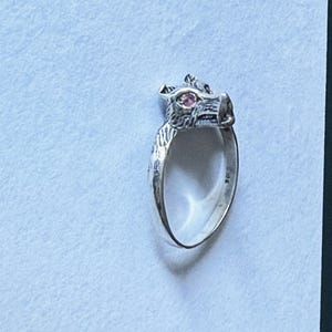 May include: A silver ring featuring a wolf head design with a pink gemstone eye. The ring is detailed with textured fur and a stylized wolf face. The ring is set against a light background.