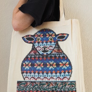 Puede incluir: Una bolsa de lona blanca con un dise&ntilde;o de oveja estampada en azul, rojo y blanco. El texto "SCOTLAND" est&aacute; impreso en verde debajo de la oveja.