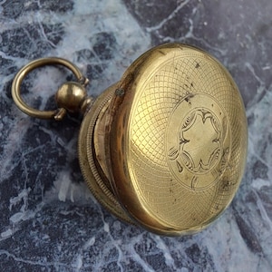 May include: A gold-toned pocket watch with a decorative, engraved design on the lid. The watch has a hinged lid and a ring for attaching a chain.