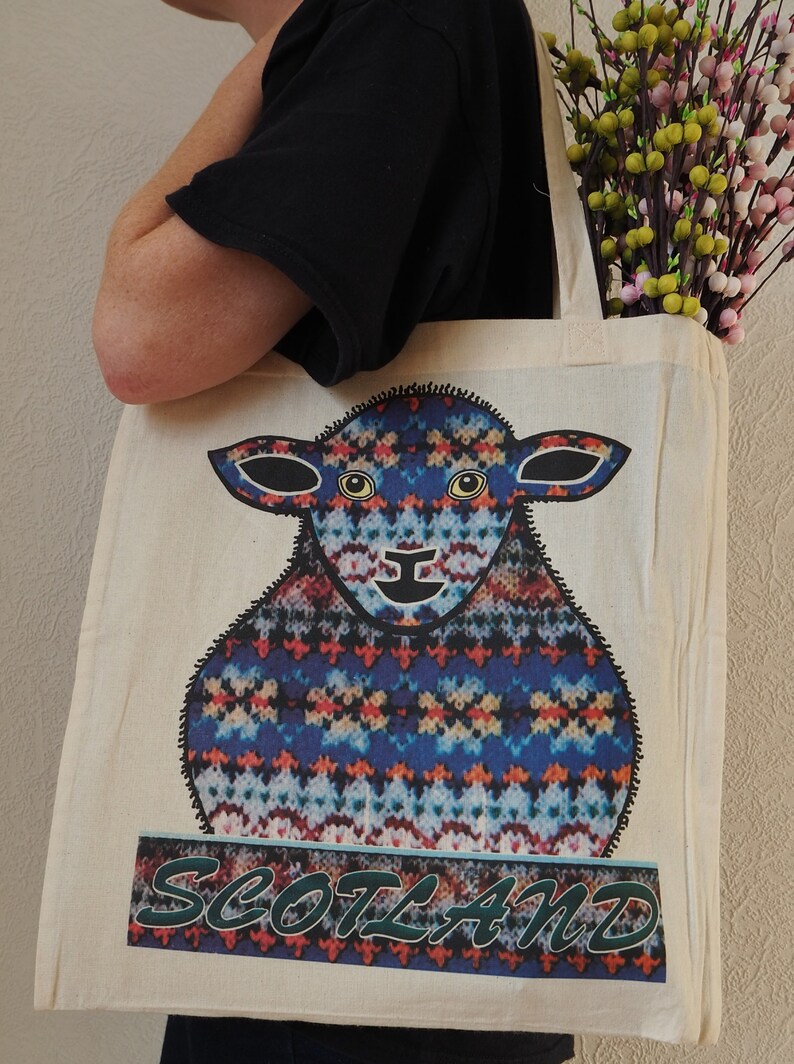 Puede incluir: Una bolsa de lona blanca con un dise&ntilde;o de oveja estampada en azul, rojo y amarillo. La bolsa tiene un texto "SCOTLAND" verde y blanco debajo de la oveja.