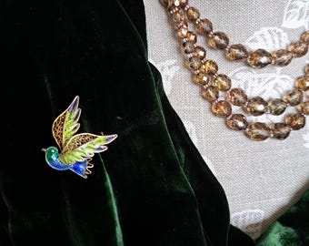 以下が含まれることがあります： 緑色のベルベット生地に、青、緑、金の鳥のブローチが付いている。鳥は羽を広げて飛んでいる。生地は、茶色のビーズネックレスをしたマネキンにかけられている。