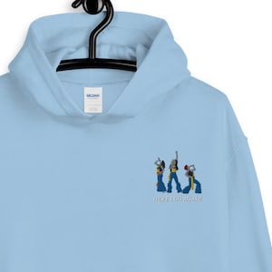 May include: Light blue hoodie with a front pocket and a drawstring hood. The hoodie features an embroidered design of three figures and the text "HERE WE GO AGAIN" in blue thread. The hoodie is hanging on a black hanger.