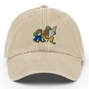 Könnte beinhalten: Beige Baseballkappe mit einem gestickten Design von zwei Fröschen. Ein Frosch trägt eine blaue Hose, der andere eine gelbe Hose. Die Frösche halten ein braunes Objekt und ein weißes und graues Objekt.