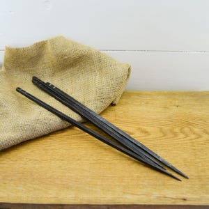 Peut inclure: Un ensemble de quatre baguettes en métal foncé posées sur une surface en bois, à côté d'un sac en toile de jute. Les baguettes sont fines et ont une forme carrée en haut. En arrière-plan, un mur en bois blanc.