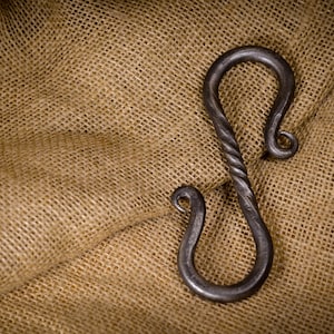 Peut inclure: Un grand crochet en métal noir forgé à la main avec un design torsadé. Le crochet est posé sur un tissu de jute brun.