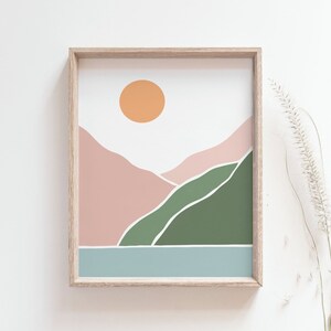 May include: A framed art print featuring a minimalist landscape design. The artwork showcases a peach-colored sun, pink and green hills, and a blue body of water. The print is encased in a light brown wooden frame, suitable for home decor.