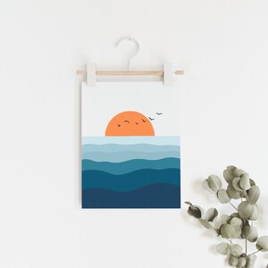 Op de afbeelding: Een digitale print van een zonsondergang boven de oceaan met een gestileerde oranje zon en blauwe golven. De print hangt aan een witte muur met een houten lat en een witte haak.