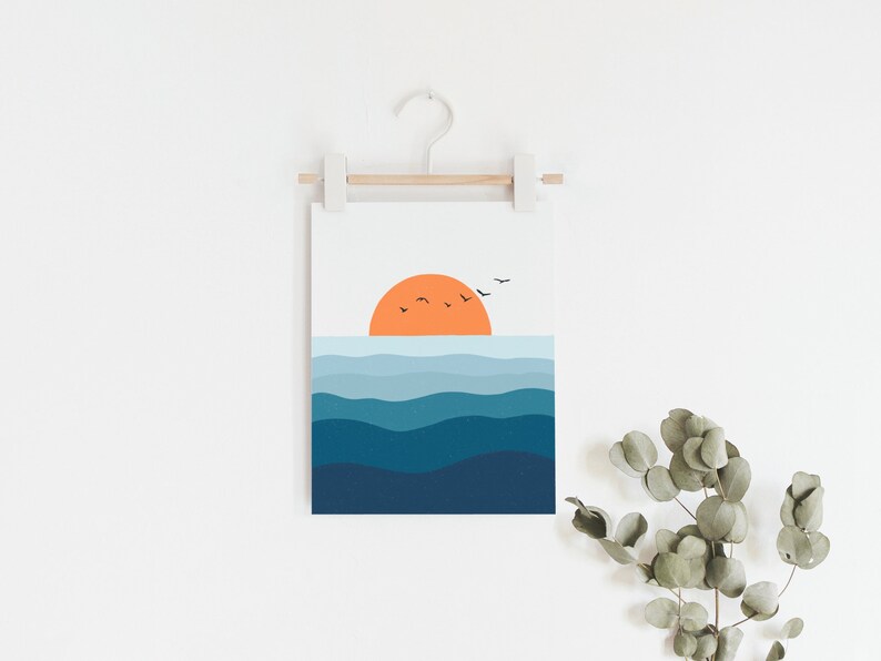 Op de afbeelding: Een digitale print van een zonsondergang boven de oceaan met een gestileerde oranje zon en blauwe golven. De print hangt aan een witte muur met een houten lat en een witte haak.