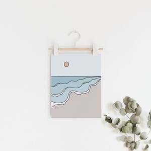 May include: A minimalist line art print of a beach scene with blue waves and a setting sun. The print is hanging on a white wall with a wooden dowel and white clips.