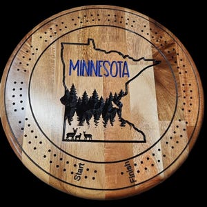 May include: A wooden game board shaped like the state of Minnesota with a black forest design and deer silhouettes. The board has a circular shape with a black outer ring and a dotted pattern. The words "Start" and "Finish" are written on the board.