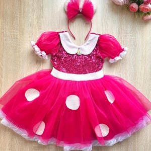 May include: A pink and white tulle dress with white polka dots and a sequined bodice. The dress has a white collar and white lace trim. A pink tulle headband with a bow and a rhinestone charm is included.