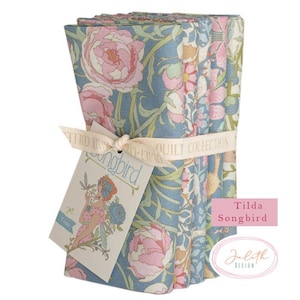 Könnte beinhalten: Ein Stapel quadratischer Stoffstücke mit Blumenmuster, gebunden mit einem cremefarbenen Band. Der Stoff hat einen blauen Hintergrund mit rosa Rosen und grünen Blättern. Ein Etikett mit dem Wort "Songbird" und einer floralen Illustration ist angebracht. Der Text "Tilda Songbird" ist ebenfalls sichtbar.