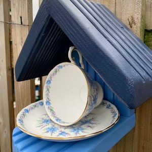 May include: A blue birdhouse with a white teacup and saucer. The teacup and saucer have a floral pattern with blue flowers and gold trim.
