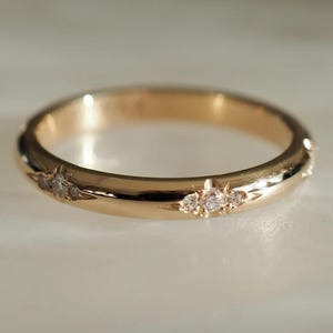 Op de afbeelding: Een gouden ring met kleine, sprankelende edelstenen. De ring heeft een glad, gepolijst oppervlak en een klassiek bandontwerp. De edelstenen zijn in kleine groepen rond de omtrek van de ring gegroepeerd.