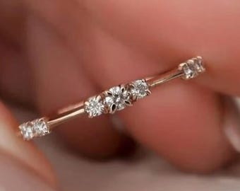 Könnte beinhalten: Ein zarter Goldring mit einer Reihe kleiner, funkelnder Diamanten. Der Ring hat ein dünnes Band und ist mit mehreren rund geschliffenen Diamanten besetzt, wodurch ein subtiles, aber elegantes Design entsteht. Der Ring wird von einer Person gehalten.