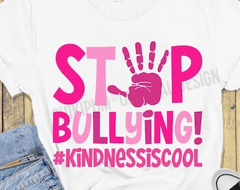 May include: A white t-shirt with a pink handprint and the text "STOP BULLYING! #KINDNESSISCOOL" in pink letters. The shirt is on a wooden background with a pair of black sneakers and a pair of blue jeans.