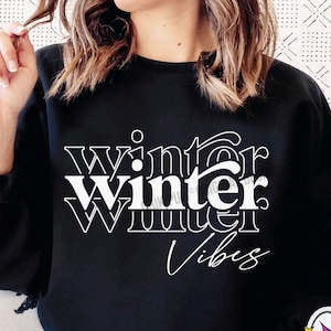 Puede incluir: Una sudadera negra con el texto "winter Winter Vibes" en blanco. La sudadera la lleva puesta una persona.