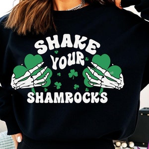 Puede incluir: Sudadera negra con un gráfico blanco que dice "Shake Your Shamrocks" con dos tréboles verdes que sostienen manos esqueléticas.
