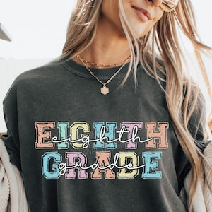 May include: A dark grey t-shirt with the text "EIGHTH GRADE" in colorful letters. The letters are outlined in white and have a distressed look. The text is centered on the shirt.