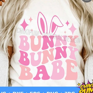 Może przedstawiać: Biała koszulka z nadrukiem różowych uszu króliczka i tekstem "BUNNY BUNNY BABE" w kolorze różowym, z nakładającymi się literami. Tekst jest w retro stylu czcionki.