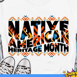 May include: A white t-shirt with a black and orange design that says "Native American Heritage Month". The design is made up of geometric shapes and patterns. The t-shirt is on a white background.