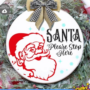 Op de afbeelding: Een wit rond bord met een rood kerstman-gezicht en de tekst "SANTA Please Stop Here" in zwart. Het bord is versierd met een zwart-wit geblokte strik en blauwe sneeuwvlokken.